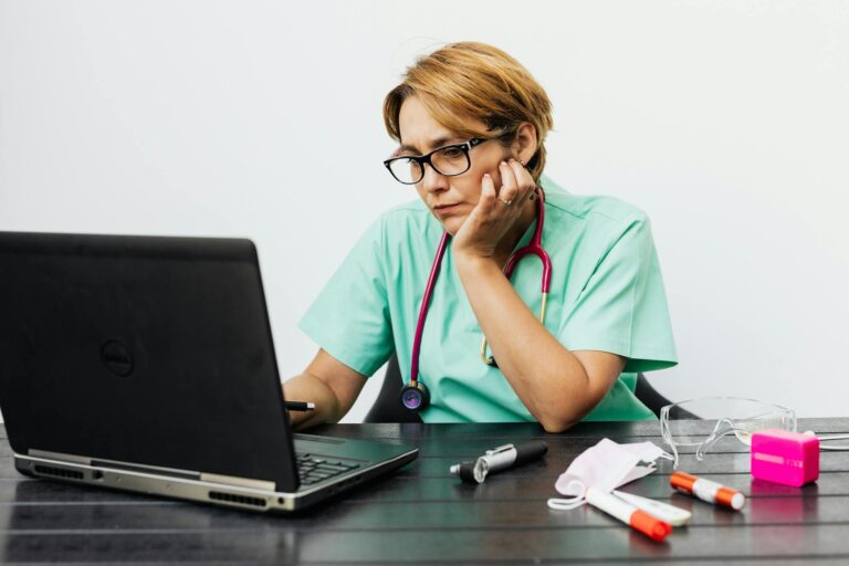 Focused nurse with eyeglasses working on a laptop surrounded by medical tools on a desk.